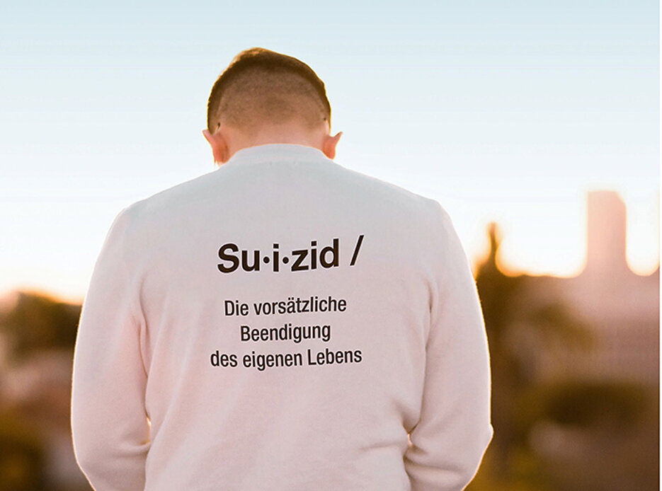 Junger Mann, von hinten fotografiert, der einen Pullover mit der Aufschrift "Suizid - die vorsätzliche Beendigung des eigenen Lebens" trägt.